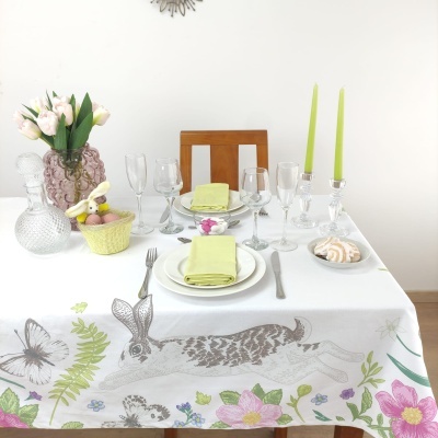 Mesa posta com toalha decorada com coelho e flores, pratos, guardanapos verdes, copos, castiçais com velas, vaso com tulipas e cesta de ovos.