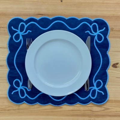 Prato branco sobre jogo americano azul com desenho em azul claro, garfo e faca, em mesa de madeira clara