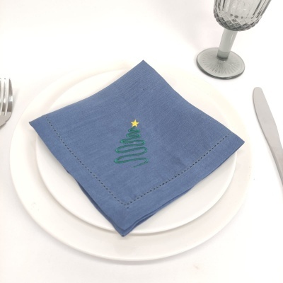 Guardanapo azul com bordado de árvore de natal colocado em pratos brancos com talheres e taça