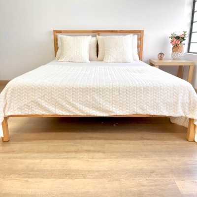 Cama de madeira com edredão branco e almofadas, mesa de cabeceira com vaso de flores e objeto redondo