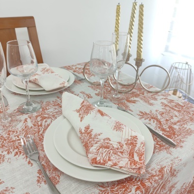 Mesa posta com toalha e guardanapo estampados em vermelho terroso, pratos brancos, copos de vidro, talheres metálicos e castiçal dourado com velas torcidas.