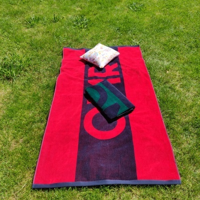 Toalha de praia vermelha e azul com letras, almofada branca colorida e toalha azul-verde sobre relva