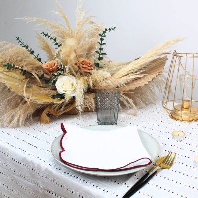 Arranjo floral com flores secas em vaso de vidro cinzento sobre mesa com toalha branca, prato, guardanapo e talheres dourados.