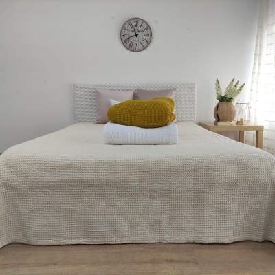 Cama com colcha tricot creme, mantas amarelo e branco dobradas, almofada lilás, cabeceira tricot, relógio cinzento, mesa com vaso e flores, cortina branca