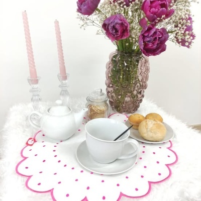 mesa decorada com flores, velas, bule, chávena e pãezinhos sobre toalha branca com pontos cor-de-rosa