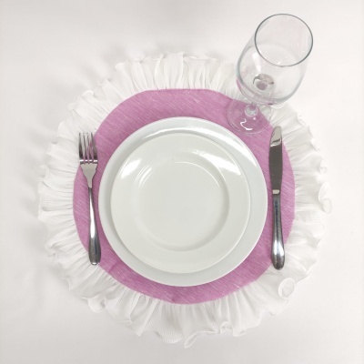 Mesa posta com prato fundo e raso brancos, talheres de metal, copo de vidro e jogo americano rosa e branco frisado.