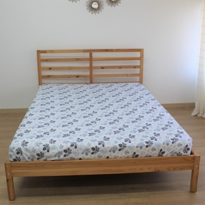 Cama de madeira clara com colchão revestido por lençol branco com flores cinzentas e pretas