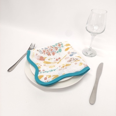 Guardanapo colorido com borda azul sobre prato branco, talheres e copo de vidro