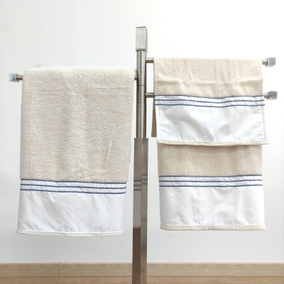 Conjunto de toalhas bege com barra branca e linhas azuis penduradas num suporte metálico.