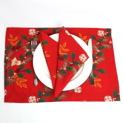 Jogo americano e guardanapo vermelhos com padrão de folhas e bolas decorativas sobre prato branco e talheres.