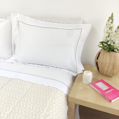 Almofada branca com borda sobre cama e mesa de cabeceira com vaso, vela e livro rosa