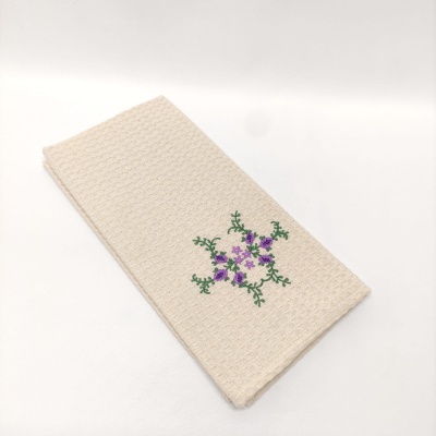 Toalha bege com bordado floral roxo e folhas verdes em fundo branco