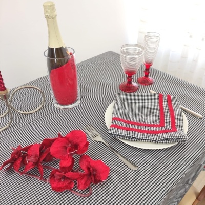 Mesa posta com toalha xadrez preto e branco, guardanapo com bordas vermelhas, talheres, taças com pés vermelhos, garrafa de champanhe em balde de vidro com capa vermelha e flores vermelhas.