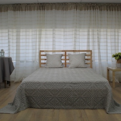 Quarto com cama de madeira clara, colcha cinza com padrão geométrico e almofadas brancas