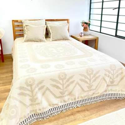 Cama com colcha bege bordada e duas almofadas, ao lado mesa de madeira com flores