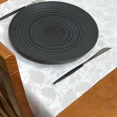 Prato preto texturizado com talheres pretos numa mesa de madeira com toalha branca floral