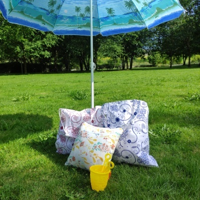 Três almofadas coloridas na relva sob um guarda-sol azul com desenhos, com balde amarelo de brinquedo para praia