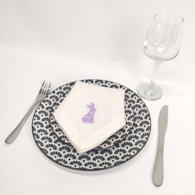 Conjunto de refeição com prato cerâmico preto e branco, guardanapo branco com coelho roxo, talheres de metal e copo de vidro.