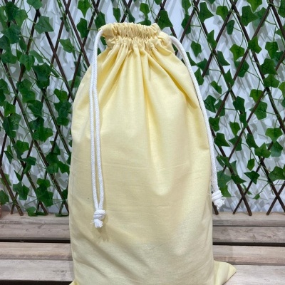 Saco de tecido amarelo claro com cordão branco, sobre madeira, com fundo de treliça e folhas verdes