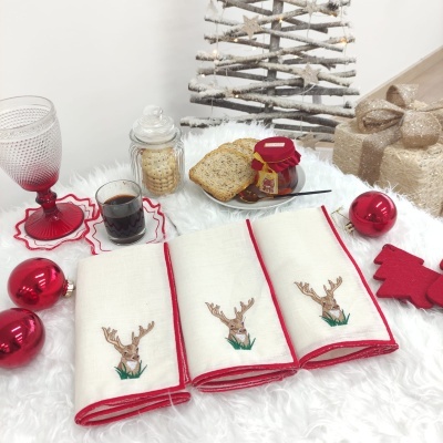 Três toalhas com bordado de veado e bordas vermelhas em mesa com decoração natalícia e alimentos