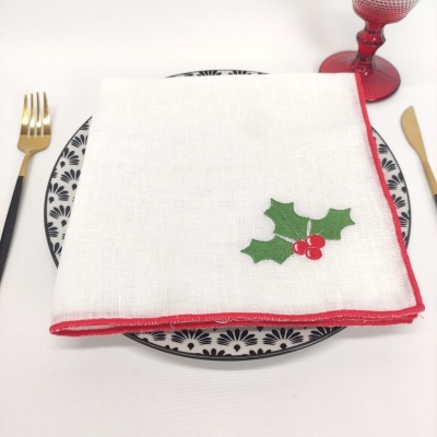 Guardanapo branco com borda vermelha e decoração de folhas verdes e bagas vermelhas sobre prato preto e branco com padrão floral