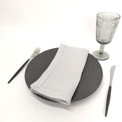 Conjunto de louça com prato preto, guardanapo cinza, talheres pretos e copo cinzento em superfície branca