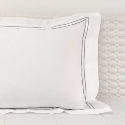Almofada branca com bordadura cinza sobre lençol branco e almofada texturizada branca