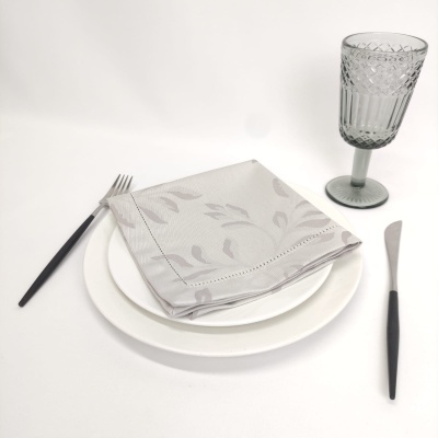 Mesa posta com talheres pretos, prato branco, guardanapo cinzento com padrão de folhas e copo de vidro cinzento