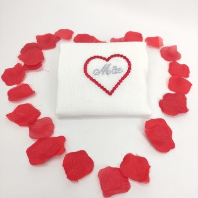 Toalha branca com bordado de coração e palavra 'Mãe' rodeada por pétalas de rosa vermelhas