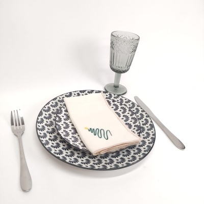 Conjunto de louça com pratos azuis, guardanapo branco bordado, talheres prateados e copo de vidro com textura