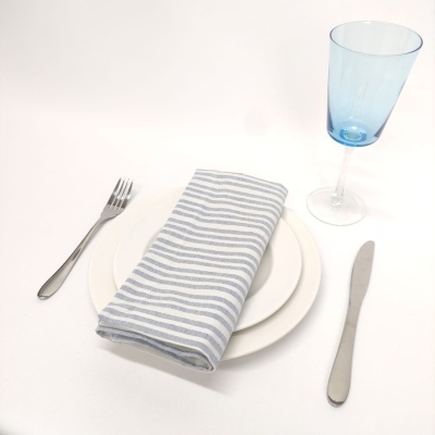 Prato branco com guardanapo riscado cinza e branco, talheres metálicos e copo azul claro sobre mesa branca
