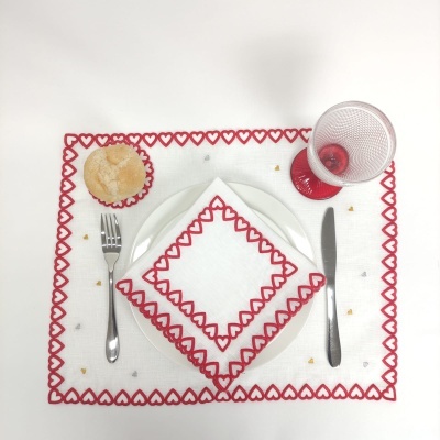 Conjunto de mesa com individual e guardanapo branco com bordas vermelhas em forma de corações, copo vermelho, pão, faca e garfo