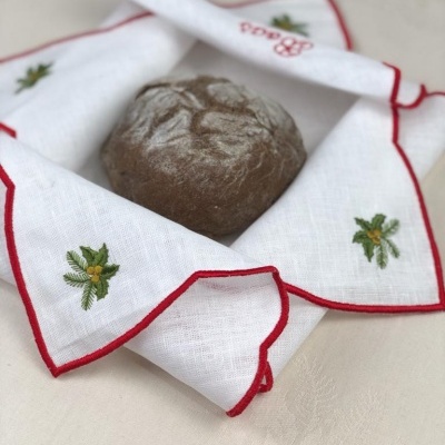 Pano de cozinha branco com bordas vermelhas e bordados de pinhas verdes envolvendo pão redondo escuro