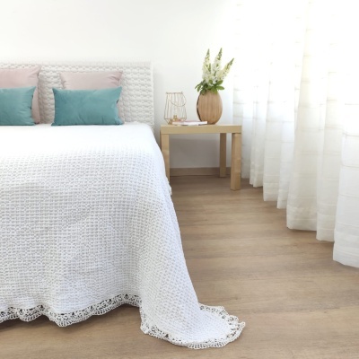 Quarto com cama com colcha branca texturizada e almofadas pastel, mesa de apoio com vaso e flores, cortinas brancas e chão de madeira clara