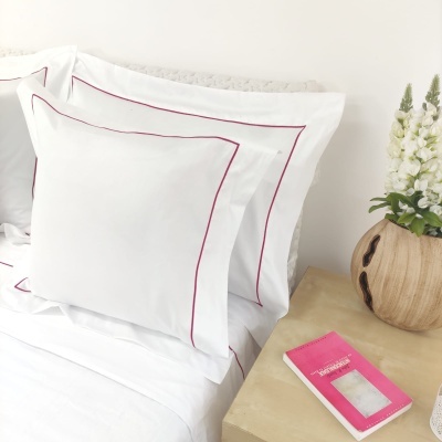 Conjunto de almofadas brancas com linhas vermelhas numa cama ao lado de vaso de flores e livro cor-de-rosa