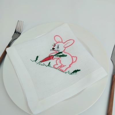 Guardanapo branco bordado com coelho cor-de-rosa segurando cenoura, sobre prato branco