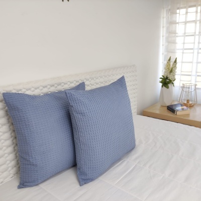 Duas almofadas azuis texturizadas em cama branca com cabeceira e mesa de cabeceira com flores