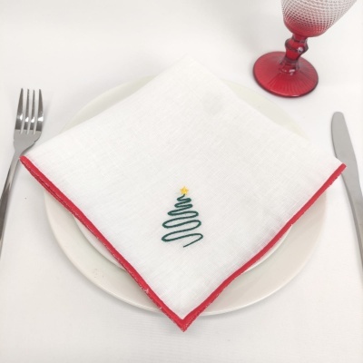 Guardanapo branco com borda vermelha e árvore de Natal bordada, prato branco, talheres e copo vermelho à mesa