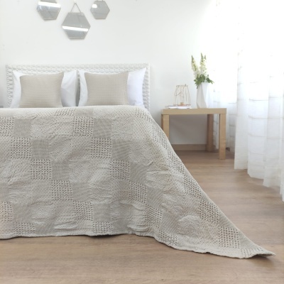 Cama com colcha cinzenta clara de croché e almofadas, mesa de cabeceira de madeira, vaso de flores e luminária, ambiente quarto claro e decorado.