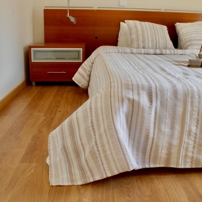 Cama com colcha branca de riscas cinzentas e almofadas, cabeceira e mesa de cabeceira em madeira castanha, chão em madeira clara