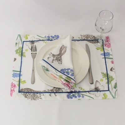 Mesa posta com prato branco, talheres de metal, copo de vidro e jogos de guardanapo e individual estampados com coelho e flores