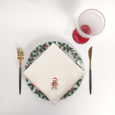 Mesa posta com prato decorado, guardanapo branco com bordado de biscoito de gengibre, talheres metálicos, e copo transparente com base e bola vermelha.