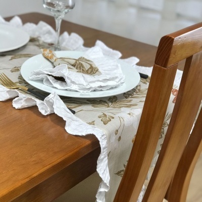 Mesa de madeira com toalha creme decorada, pratos brancos, guardanapos brancos e talheres dourados