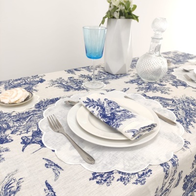 Mesa posta com toalha branca e padrão azul, prato branco, talheres, copo azul, decantador e vaso com flores.