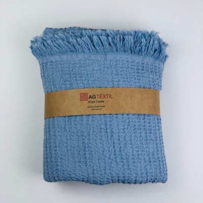 Manta azul com padrão de pequenos quadrados e franjas, embrulhada com faixa de papel castanho com texto AG TÊXTIL