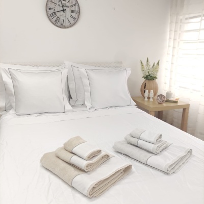 Cama com roupa de cama branca, pilhas de toalhas bege e branco, mesa de cabeceira com vasos, relógio de parede e janela com cortina