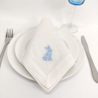 Guardanapo branco com bordado azul de coelho sobre prato branco, talheres e copo azul