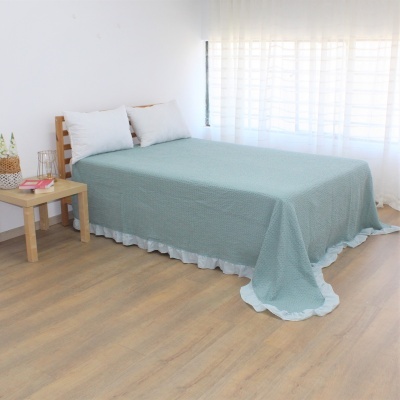 Cama com colcha verde-água e almofadas brancas junto a mesa de cabeceira e janela.