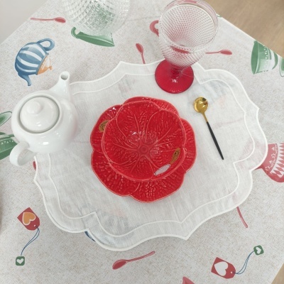 Conjunto de chá com bule branco, copos, prato vermelho em forma de flor, colher dourada e toalha decorada com bules coloridos.