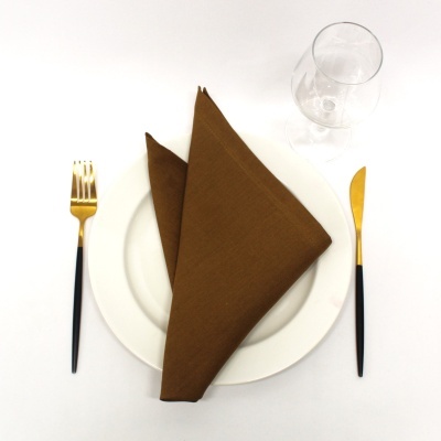 Conjunto de jantar com prato branco, guardanapo castanho, talheres dourados e copo transparente